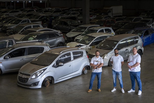 Three car buyers standing in an indoor warehouse with a silver Chevrolet Spark missing a front wheel.